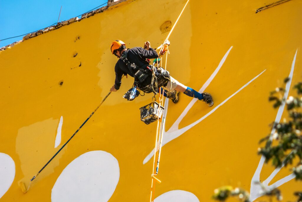 A man rappelling down a building, creating vibrant street art on a yellow wall.